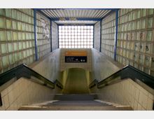 Treppenabgang zu einem unterirdischen Durchgang mit vergitterten Glasbausteinwänden und metallischem Dach. Im Hintergrund befindet sich ein Hinweisschild mit Informationen zu Zügen und Durchgangsrichtungen.