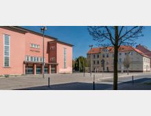 Außenansicht eines rosa Gebäudes mit der Aufschrift „Stadttheater“, flankiert von hohen Fenstern und einem flachen Dach; davor ein gepflasterter Platz mit Straßenlaternen und einem kahlen Baum. Im Hintergrund befinden sich Wohnhäuser mit roten Ziegeldächern und beige Fassaden.