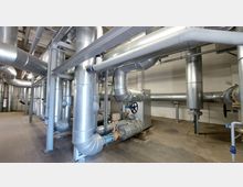 Industrieanlage mit zahlreichen silberfarbenen Rohren und Leitungen, die an der Decke und an Metallträgern befestigt sind. Im Vordergrund befindet sich ein großes Radventil sowie weitere Steuerungselemente, umgeben von einem offenen Raum mit Betonboden und einer hellen Wand.