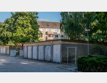 Mehrere Garagen mit verwitterten, weißen Toren stehen nebeneinander auf einem gepflasterten Hof. Im Hintergrund sind Bäume und ein mehrstöckiges Wohnhaus mit Balkonen zu sehen.