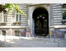 Alttext: Ehemaliges Wohngebäude mit verwitterter Fassade, hohen Fenstern und einem großen Eingangsportal mit einem Rundbogen, flankiert von verzierten Säulen. Die Fassade ist mit Graffiti bedeckt, und der Eingangsbereich zeigt abgeblätterte Farbe und Holzdetails.