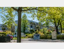 Mehrfamilienhaus mit moderner Architektur, umgeben von dichter Begrünung und Bäumen. Im Vordergrund befinden sich eine Straße, geparkte Autos, ein Stromkasten mit Graffiti und ein Fußweg.