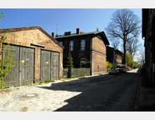 Straßenansicht in einem historischen Viertel mit gepflastertem Weg, umgeben von Ziegelgebäuden. Links befindet sich ein Nebengebäude mit großen Holztoren, rechts stehen Wohnhäuser und vereinzelte Bäume entlang der Straße.