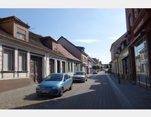 Eine schmale Kopfsteinpflasterstraße in einer Kleinstadt, gesäumt von niedrigen historischen Gebäuden mit Schaufenstern und Schildern, darunter ein Schneideratelier. Am Straßenrand parken mehrere Autos, und im Hintergrund sind Bäume und weitere Häuser zu sehen.