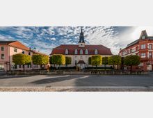 Historisches Gebäude mit rotem Mansardendach und einem zentralen Uhrturm, umgeben von symmetrisch angeordneten, gestutzten Bäumen. Das Gebäude steht zwischen zwei mehrstöckigen Häusern und befindet sich an einer gepflasterten Straße bei sonnigem Wetter mit teils bewölktem Himmel.