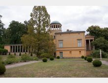 Historisches Gebäude mit gelber Fassade, einer runden Kuppel und einem Säulengang, umgeben von Bäumen und einer gepflegten Gartenanlage mit Wegen und Sträuchern. Im Vordergrund steht eine weiße Büstenstatue auf einem Sockel.