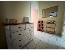 Heller Eingangs- oder Flurbereich mit weiß gefliestem Boden und cremefarbenen Wänden, ausgestattet mit einer weißen Kommode und einem offenen Durchgang zu einem Nebenraum. An der Wand hängen ein großer rechteckiger Spiegel; daneben stehen ein terrakottafarbenes Regal mit runden Fächern sowie ein kleines Holzregal mit Dekoration.