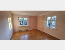 Helles, leeres Zimmer mit Holzdielenboden, cremefarbenen und pastell-rosa gestrichenen Wänden, sowie zwei Fenstern, die einen Ausblick auf eine baumreiche Umgebung bieten. Unter einem Fenster befindet sich ein Heizkörper mit einer kleinen Tischlampe.