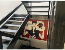 Treppenhaus mit modernen, dunklen Holztreppen und einer Glasbrüstung. An der Wand hängt ein großflächiges Pop-Art-Gemälde mit einer Frau, die eine Zeichnung mit dem Wort "POW!" in der Hand hält.