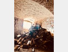 Ein rustikaler Kellerraum mit gewölbter Ziegeldecke und unverputzten Backsteinwänden, in dem ein großer Haufen gespaltenes Brennholz auf dem Boden gestapelt ist. Durch ein kleines Fenster mit Gitter dringt natürliches Licht ein.