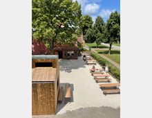 Ein Außengelände mit Kiesboden, auf dem mehrere Biertische und -bänke stehen. Im Hintergrund befinden sich ein kleines Gebäude mit roten Dachziegeln und pinker Fassade sowie große grüne Bäume; links im Vordergrund ist ein kleines Gebäude aus Holz sichtbar.