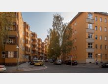 Straßenszene in einem Wohngebiet mit orangefarbenen, mehrstöckigen Apartmentgebäuden im Stil der 1920er bis 1930er Jahre. Auf der gepflasterten Straße stehen geparkte Autos, während Bäume den Straßenrand säumen.