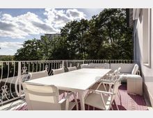 Terrasse mit weißem Tisch, mehreren weißen Stühlen und einer weißen Sitzbank, umgeben von einem dekorativen Geländer. Im Hintergrund sind Bäume und ein Gebäude unter leicht bewölktem Himmel zu sehen.