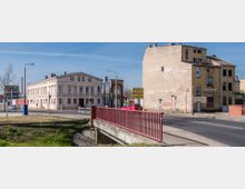 Straßenszene mit einem historischen weißen Gebäude mit Säulen und Giebel links und einem teilweise verfallenen Gebäude aus gelbem Backstein rechts. Im Vordergrund eine kleine Brücke mit rotem Geländer, und im Hintergrund Verkehrsampeln und Straßenschilder.