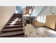 Treppe in einem älteren Gebäude mit braunen Holzstufen und einem Metallgeländer. Der Bereich bietet eine Plattform mit hellen Fliesen und einem großen Fenster, durch das Tageslicht einfällt, sowie sichtbare Rohrleitungen an der Rückwand.