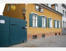 Gelbes Gebäude mit grünen Fensterläden, einem Schild mit der Aufschrift „H. Riel“ und einem Pferdekopf-Emblem. Links befindet sich ein großes, grün gestrichenes Holztor mit einem Schild „Ausfahrt Tag u Nacht freihalten“ und einer Straßenlaterne darüber.