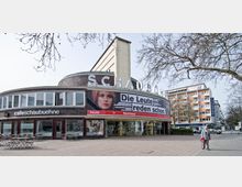 Außenansicht eines modernen Rundgebäudes mit der Aufschrift „Schaubühne“ und großer Plakatwerbung auf der Fassade. Im unteren Bereich befindet sich ein Café mit der Aufschrift „cafeschaubuehne“, davor stehen mehrere Tische und Stühle im Außenbereich; daneben Gehweg mit einem einzelnen Passanten.