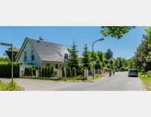 Eine ruhige Wohnstraße mit einem modernen Einfamilienhaus mit Satteldach und Garten links, umgeben von grüner Vegetation. Auf der Straße fahren zwei Radfahrer, während ein Auto am rechten Straßenrand parkt; Verkehrsschilder und Laternenpfähle sind ebenfalls zu sehen.