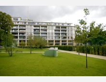 Moderner Wohnblock mit großen Glasfenstern und Balkonen, umgeben von einem gepflegten Grünbereich mit Rasen, Bäumen und einem kleinen Weg. Im Vordergrund befindet sich ein abstraktes grünes Kunstobjekt.