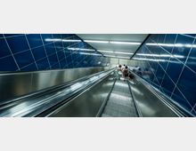 Eine Rolltreppe in einer U-Bahn-Station, umgeben von dunkelblau gefliesten Wänden und Metallgeländern. Drei Personen bewegen sich nach unten in Richtung des Bahnsteigs, der am Ende der Rolltreppe sichtbar ist.
