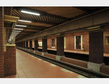 U-Bahn-Station mit Backsteinwänden, säulenartiger Architektur und einem Fliesenboden. Die Decke ist mit braunen Lamellen versehen, und entlang der Wände sind Gemälde angebracht; das Gleis und der Bahnsteig verlaufen parallel.