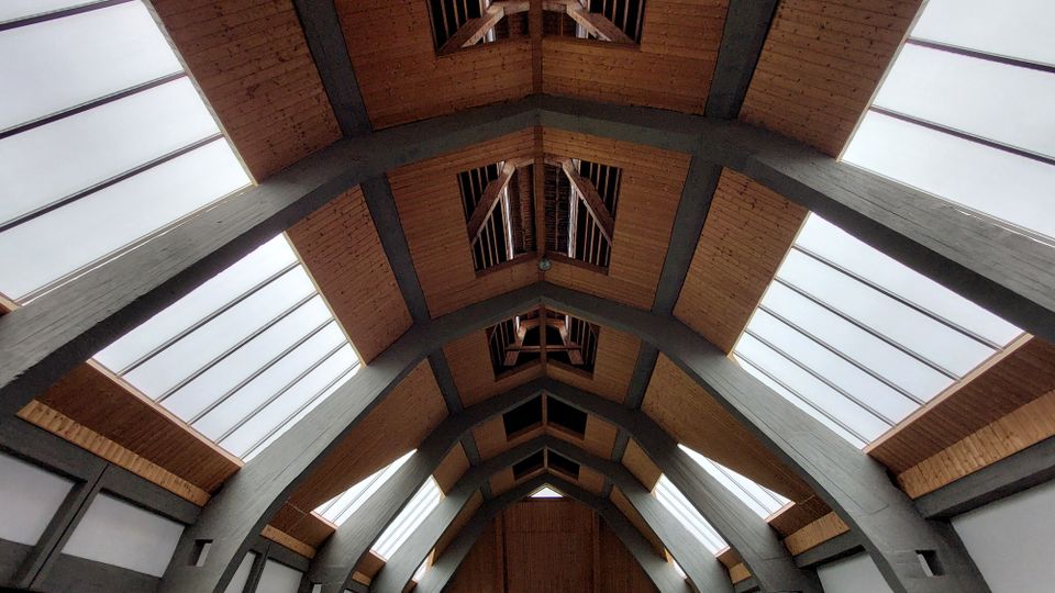 Dachansicht eines modernen Gebäudes mit einer Holzkonstruktion und großen, schrägen Glasflächen, die von dunkelgrauen Stahlträgern gestützt werden. Die Gestaltung zeigt eine symmetrische Anordnung mit geometrischen Formen und natürlichem Licht.