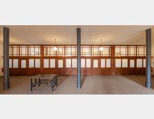 Großer Raum mit einer Wand aus hölzernen Elementen und matten Glaseinsätzen, unterteilt durch mehrere Türen. Zwei schwarze Säulen und ein schmiedeeiserner Tisch stehen in der Mitte des Raumes, der von Kugellampen an der Decke beleuchtet wird.