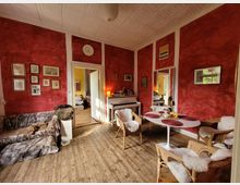 Wohnzimmer mit roten Wänden, einem Holzboden und dekorativen Bildern an den Wänden. Im Raum befinden sich ein Sofa mit Kunstfell-Decke, ein weißer Esstisch mit geflochtenen Stühlen sowie ein Klavier; durch eine geöffnete Tür ist ein angrenzendes Zimmer sichtbar.