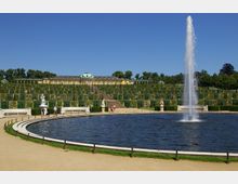 Großer Garten mit einem runden Brunnen im Vordergrund, aus dem ein hoher Wasserstrahl aufsteigt, umgeben von Statuen, Bänken und gepflegten Wegen; im Hintergrund ein terrassenförmiger Weinberg und das Schloss Sanssouci mit seiner gelben Fassade und Kuppel.