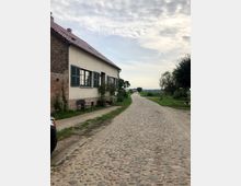 Pflasterstraße in ländlicher Umgebung mit einem historischen Gebäude aus Backstein und Putz auf der linken Seite, das grüne Fensterläden und ein rotes Ziegeldach hat. Im Hintergrund sind Wiesen, Bäume und ein leicht bewölkter Himmel zu sehen.