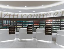 Modern gestaltete Apotheke mit geschwungenen weißen Theken im Vordergrund und einer Rückwand mit beleuchteten Regalen, die eine Vielzahl von Medikamenten und Produkten präsentieren. An der Wand darüber sind Begrüßungen in verschiedenen Sprachen zu sehen.