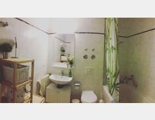 Kleines Badezimmer mit weißen Wandfliesen, einem Waschbecken mit Spiegel darüber, einer Toilette und einer Badewanne mit grün gemustertem Duschvorhang. Ein Holzregal mit Körben und Dekoration steht an der linken Seite.