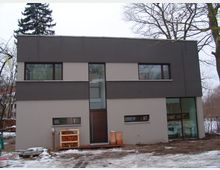 Modernes zweigeschossiges Wohnhaus mit flachem Dach und klaren geometrischen Linien. Die Fassade ist in zwei Farbtönen gehalten (dunkelgrau und beige) und verfügt über große rechteckige Fenster; im Vordergrund liegt ein schneebedeckter Garten.