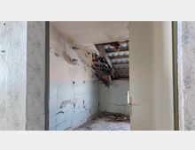 Ein verlassenes Zimmer mit beschädigter Decke, freiliegenden Dachbalken und sichtbaren Wellblechplatten. Die Wände sind rissig und teils verputzt, während der Boden mit Schutt und Holzresten bedeckt ist.