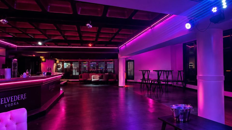 Innenraum eines modernen Club- oder Barbereichs mit dunklem Boden, langer Theke links und Sitzgruppen im Hintergrund, beleuchtet von pinken LED-Lichtleisten. Sichtbare Deckenpaneele, Stützen und hohe Stehtische entlang der rechten Wand prägen den Raum.