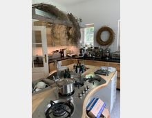 Moderne Küche mit Holzfronten und Kücheninsel, auf der sich ein Gasherd mit vier Kochfeldern, ein Spülbecken und verschiedene Utensilien befinden. An der Wand hängen getrocknete Pflanzen, ein großer Korbkranz und Regale mit Küchenmaschinen; durch ein Fenster fällt Tageslicht herein.