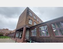 Backsteinfassade eines mehrstöckigen Gebäudes im Bauhaus-Stil mit großen, rot umrahmten Fenstern und einem überdachten Eingangsbereich. Der Himmel ist bewölkt, und im Vordergrund sind ein Gehweg aus Kopfsteinpflaster sowie ein Grünbereich erkennbar.