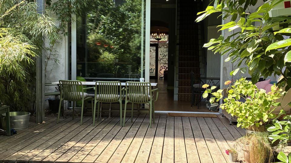 Holzterrasse mit Stufen in einem grünen Garten, mit Tisch und mehreren Stühlen vor einer großen Glasschiebetür. Die Hausfassade ist teils mit Efeu bewachsen und von dichtem Laub sowie Topfpflanzen eingerahmt.