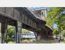 Eine erhöhte S-Bahn-Station aus Stahl mit einer Metallkonstruktion darunter, umgeben von Bäumen und einem Gehweg. Im Hintergrund verläuft ein kleiner Kanal mit einer verglasten Fußgängerbrücke darüber.