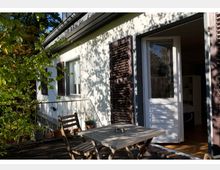 Außenansicht einer Terrasse mit Holzgeländer und Holzmöbeln, darunter ein Tisch und zwei Stühle. Das weiße Haus im Hintergrund hat weiße Fenster, braune Fensterläden und eine offene Tür, die ins Innere führt.