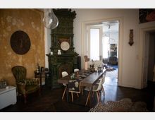 Wohnzimmer mit rustikalem Charme: Ein grüner, verzierter Kachelofen dominiert die Szene, daneben ein antiker grüner Ledersessel und ein Holztisch mit weißen Stühlen. Die Wände sind teils verputzt, teils freigelegt, und eine Tür führt in einen angrenzenden Raum mit modernen Details.