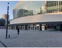 Modernes Gebäude mit geschwungener Fassade aus Glas und hellen Steinplatten, darunter ein Eingangsbereich mit großen Türen. Im Vordergrund ein gepflasterter Platz, links ein schlanker Laternenpfosten und rechts ein Geldautomat.