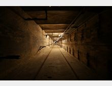 Langer, schmaler Untergrundtunnel mit gemauerten Wänden und einer sandigen Bodenfläche, durchzogen von Schienen. Die Decke ist durch Rohrleitungen und Kabelkanäle geprägt, während gleichmäßig verteilte Lampen den Raum schwach beleuchten.