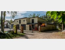 Ein Bauernhof mit gelblichem Gebäude, Tor aus Metall und Ziegelpfosten, umgeben von Bäumen und eingezäuntem Hof. Im Hintergrund sind Außenlagerbereiche und weitere Gebäude erkennbar.