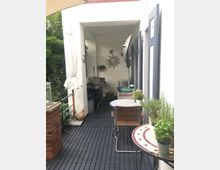 Kleiner Außenbereich mit dunklem Holzfußboden, einer gemauerten Säule links und einer überdachten Nische mit Grill und Dekorationen an der Wand. Im Vordergrund stehen zwei runde Mosaiktische mit Holzstühlen, darauf Pflanzen in Töpfen.