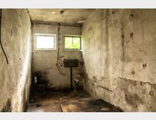 Ein kleiner, heruntergekommener Raum mit rauen, verputzten Wänden, zwei kleinen Fenstern, einem Waschbecken und sichtbaren Rohren. Der Boden ist schmutzig und voller Trümmer, während die Wände Risse und Verfärbungen aufweisen.