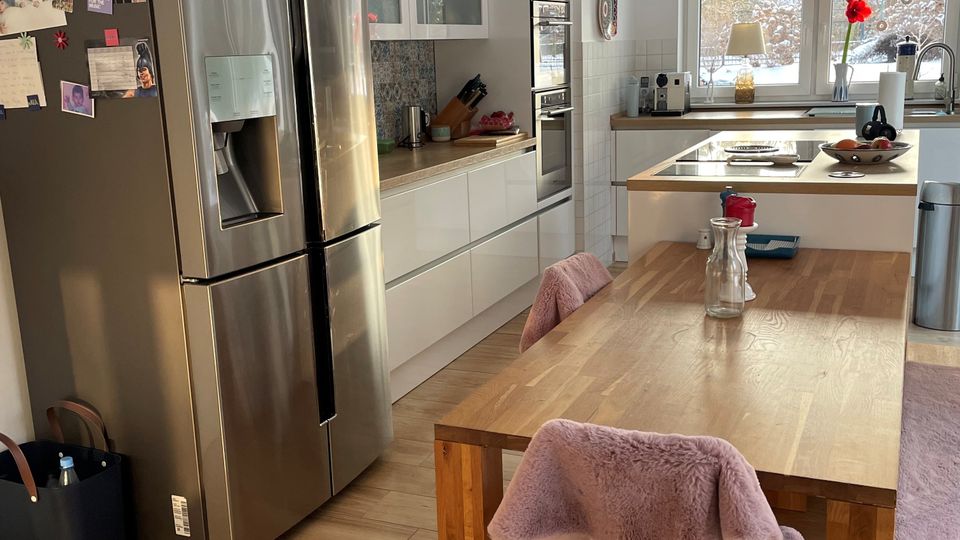 Helle, moderne Wohnküche mit weißen Einbauschränken, Edelstahl-Kühlschrank und Holzarbeitsflächen sowie großem Holzesstisch im Vordergrund. Über einer Kücheninsel und dem Tisch hängen Pendelleuchten, und ein Fenster an der Rückwand bringt Tageslicht in den Raum.