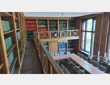 Innenansicht einer zweistöckigen Bibliothek mit holzvertäfelten Wänden, gefüllt mit Bücherregalen und farbigen Buchrücken. Im unteren Bereich befindet sich ein Besprechungstisch mit grünen Stühlen, darüber ein großer Kronleuchter, und an der Wand sind große Fenster zu sehen, die Tageslicht hereinlassen.