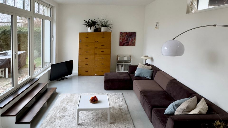 Helles, modern eingerichtetes Wohnzimmer mit hohen Decken, großflächiger Glasfront zur Terrasse und schmalen Oberlichtern an der rechten Wand. Im Raum stehen ein dunkles Ecksofa mit Bogenlampe, ein heller Teppich mit kleinem Couchtisch sowie ein hoher Holzschrank und ein niedriger Fernseher neben Stufen entlang der Fensterfront.