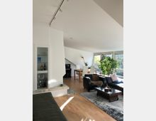 Wohnzimmer mit modernem, offenen Grundriss, ausgestattet mit hellen Holzböden, mehreren Sitzmöbeln, einem Couchtisch und einem Esstisch mit weißen Stühlen. Große Fenster lassen viel Tageslicht herein, und eine grüne Zimmerpflanze steht in der Nähe der Fenster.